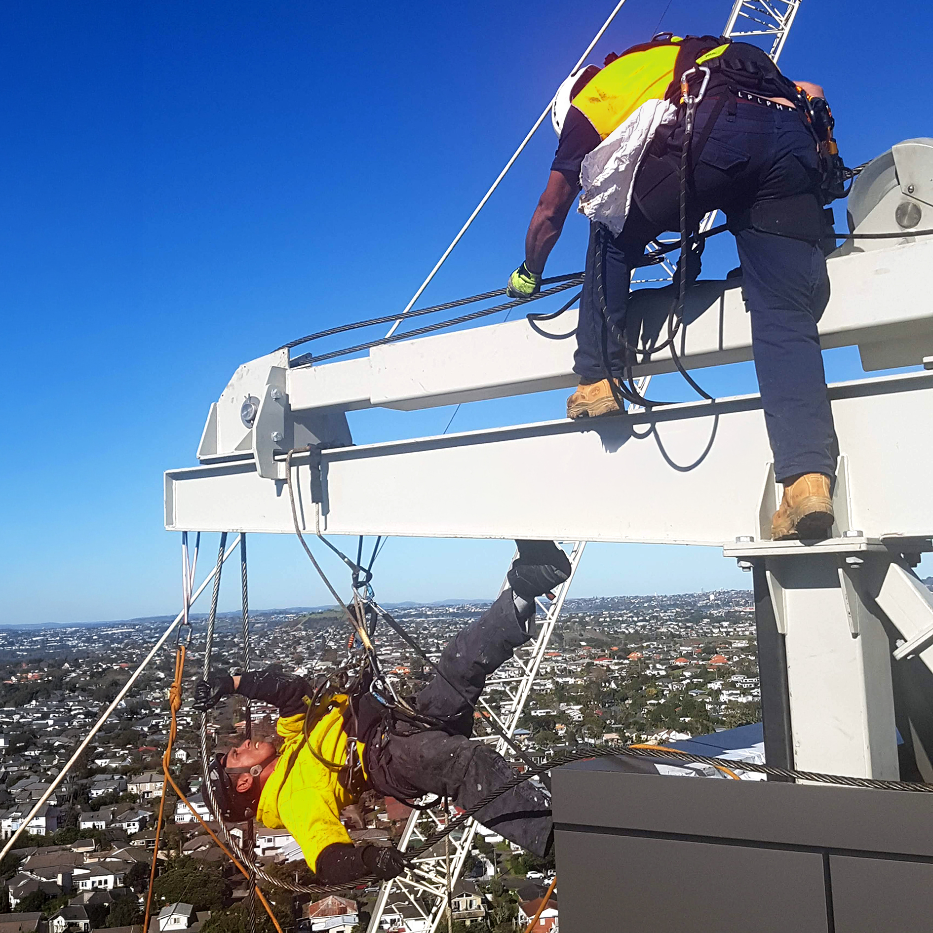 Two workers in safety gear on a tall crane structure above a cityscape on a clear day.