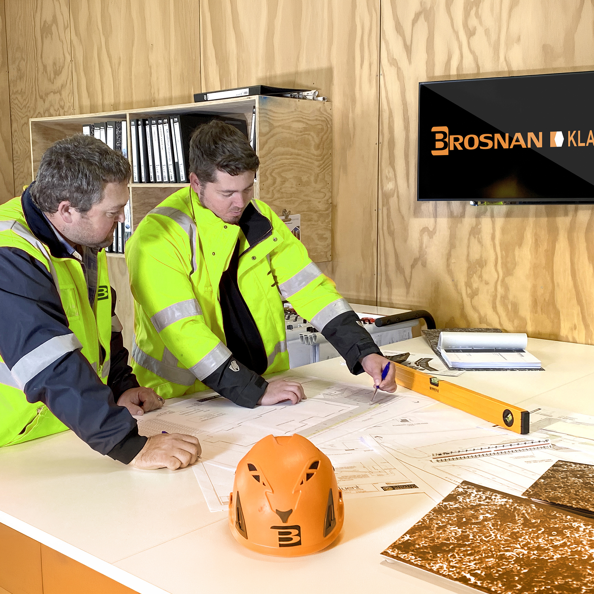 Two workers in yellow jackets reviewing building plans on a table with an orange helmet and tools.