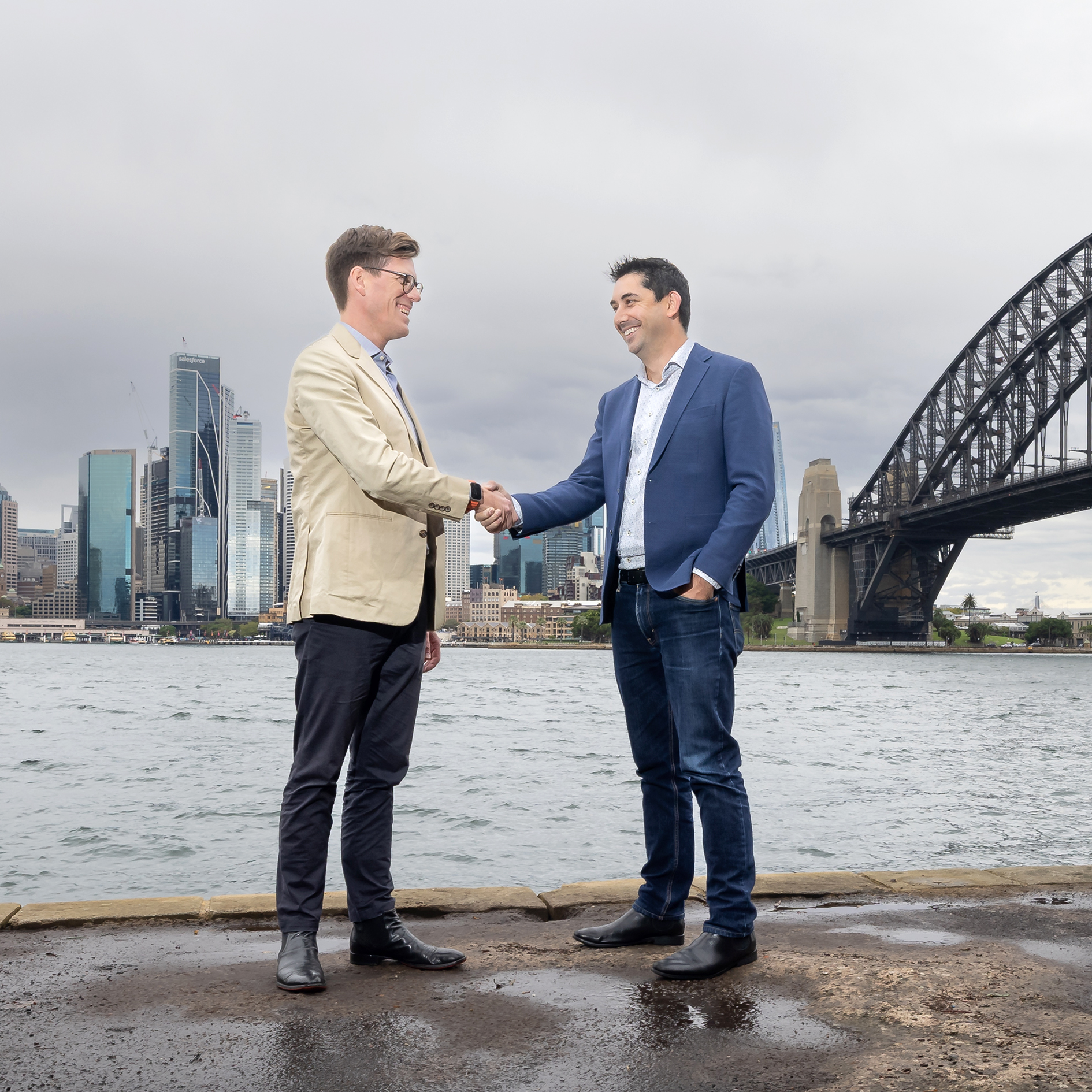 Two men in business attire shaking hands by the water with a city skyline and bridge behind them.