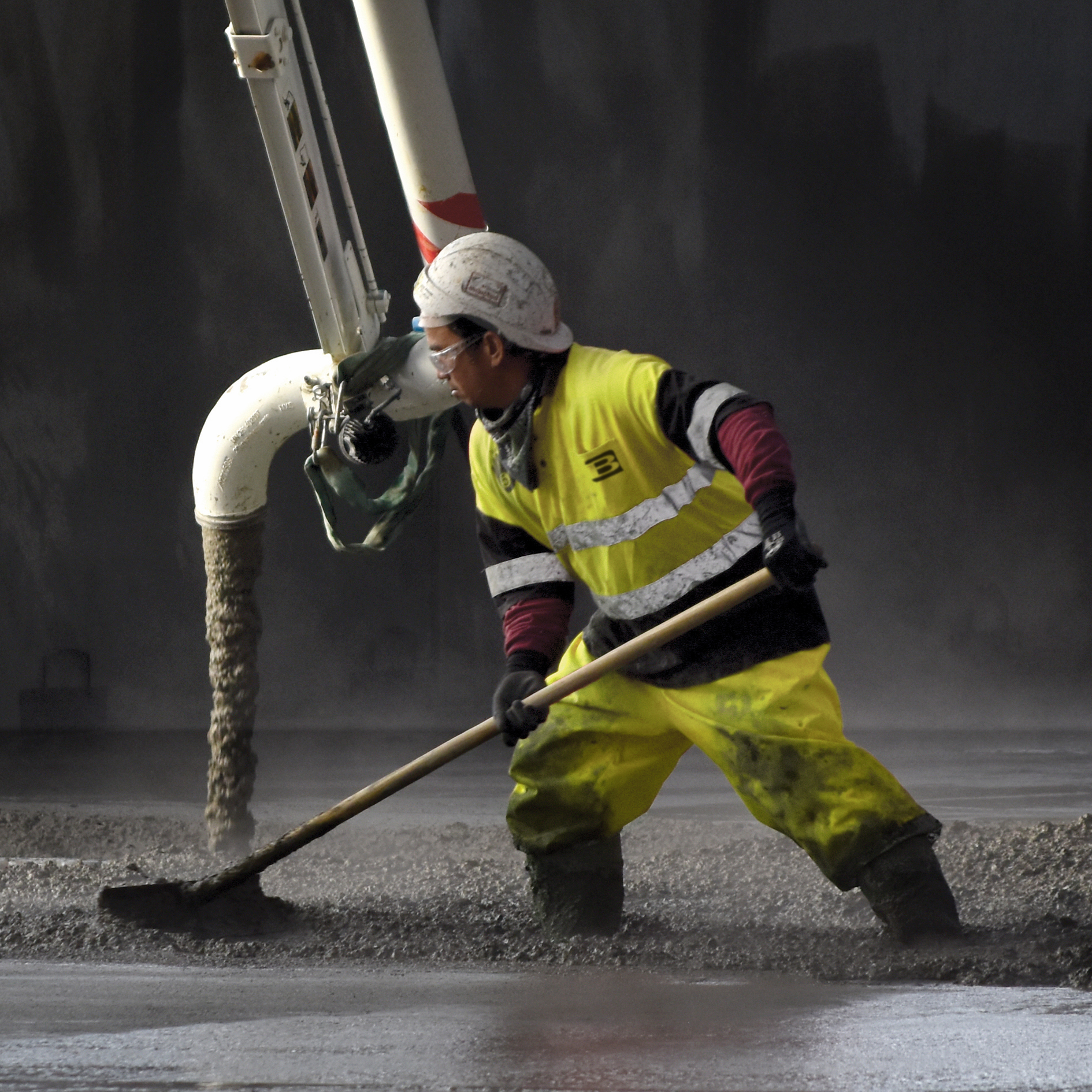 Construction worker wearing yellow gear spreading wet concrete from a large pipe on a construction site.