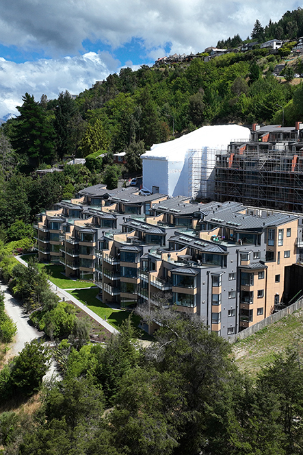 Modern apartment buildings surrounded by greenery with a hill in the background under a partly cloudy sky.