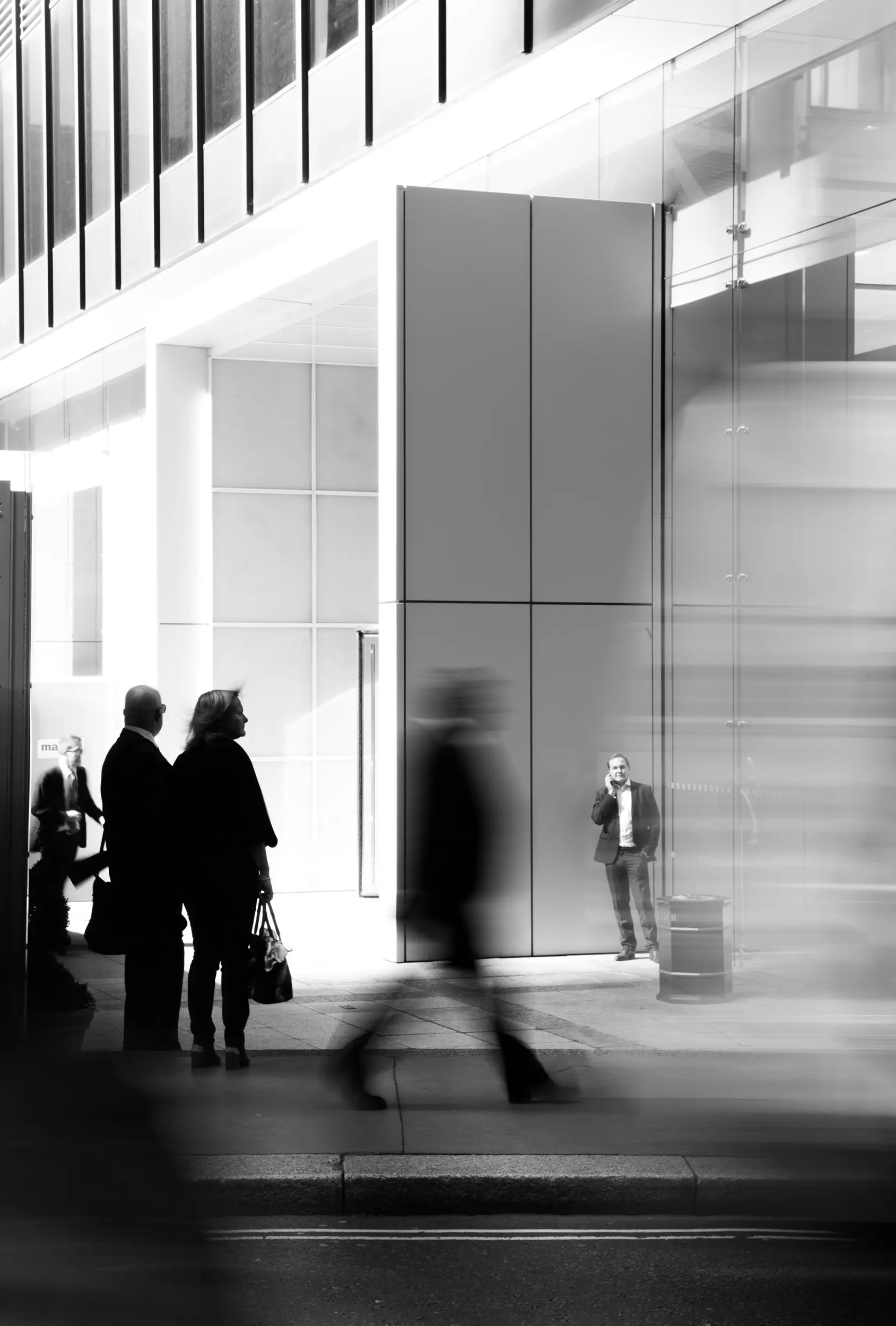 Black and white image of people standing and blurred figures walking outside a modern building.