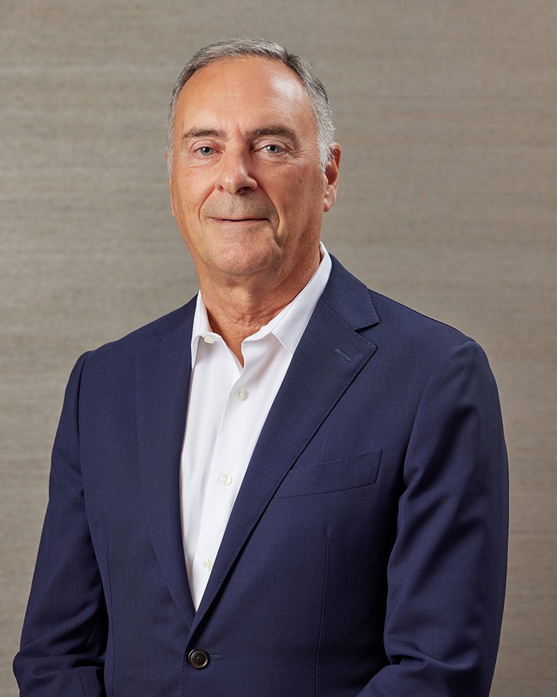 Middle-aged man wearing a navy suit jacket and white dress shirt against a neutral background.