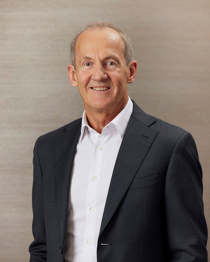 Smiling older man in a dark suit jacket and white shirt standing against a beige background.