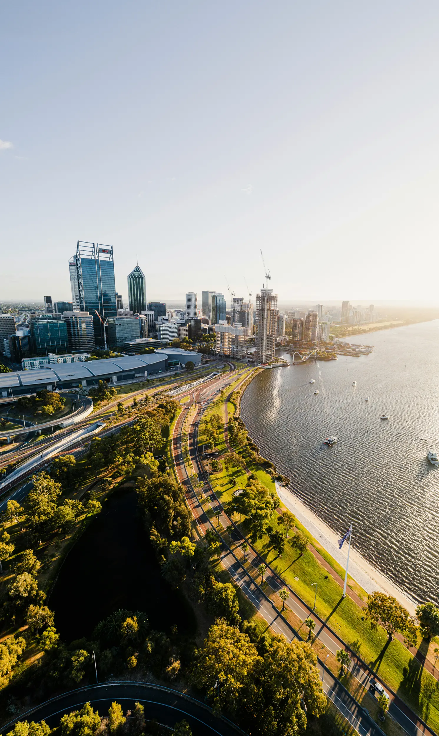 Aerial view of a city skyline by a river with boats, roads, trees, and buildings at sunset.