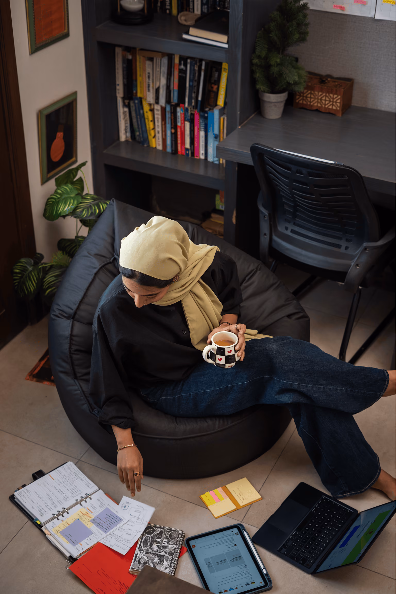 Woman in hijab sitting on a bean bag with a mug, surrounded by notebooks, a tablet, and a laptop.