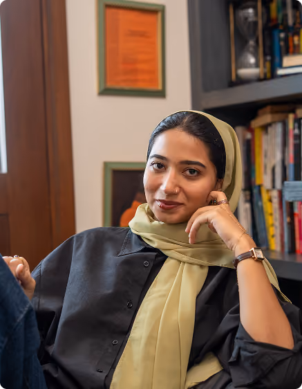 Confident woman wearing a light green hijab and black shirt sitting in a cozy room with bookshelves.