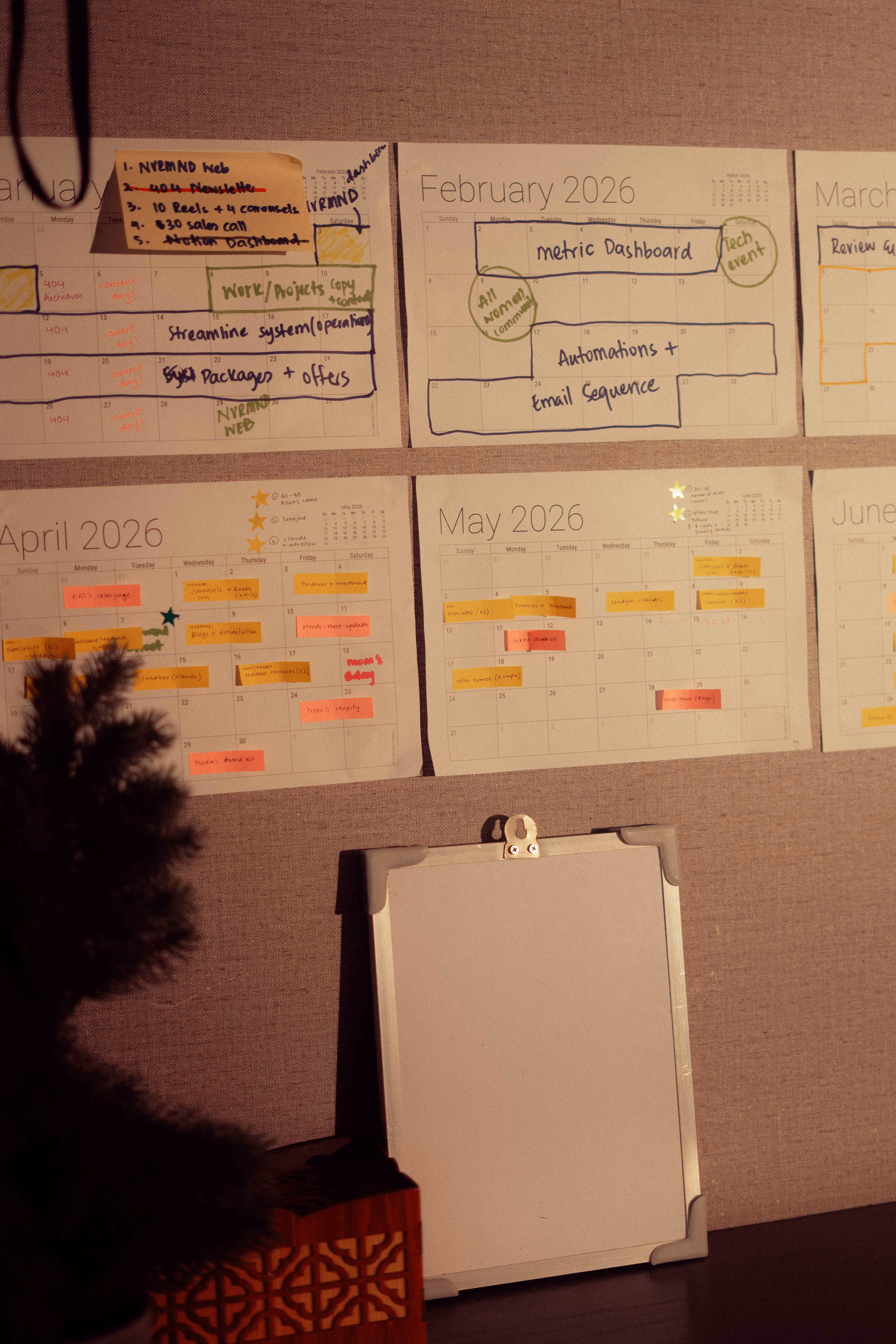 Wall calendar pages for early 2026 with handwritten notes pinned above a blank white clipboard on a table.