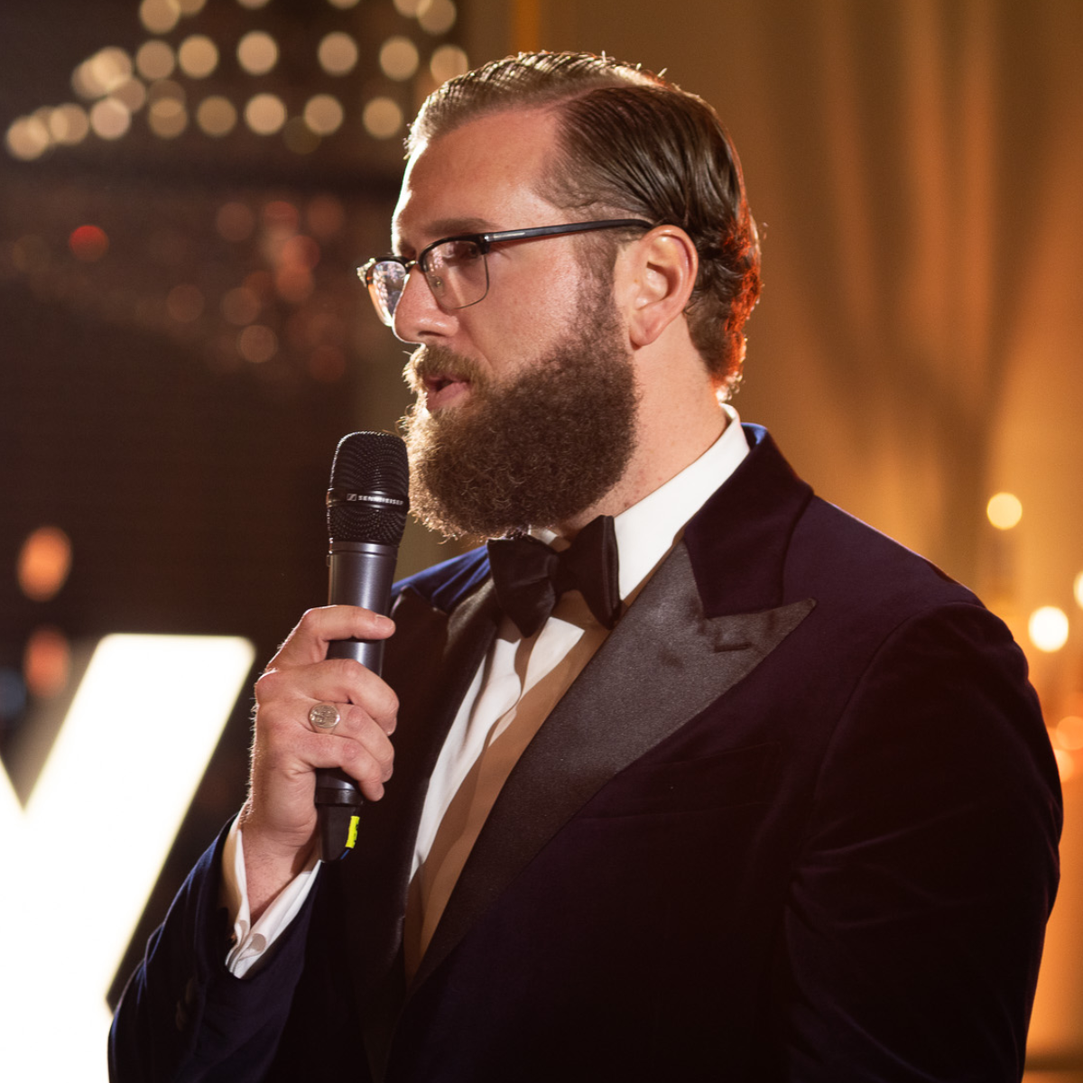 Maximilian Jäger speaking into a microphone, dressed in a black tuxedo with bow tie.