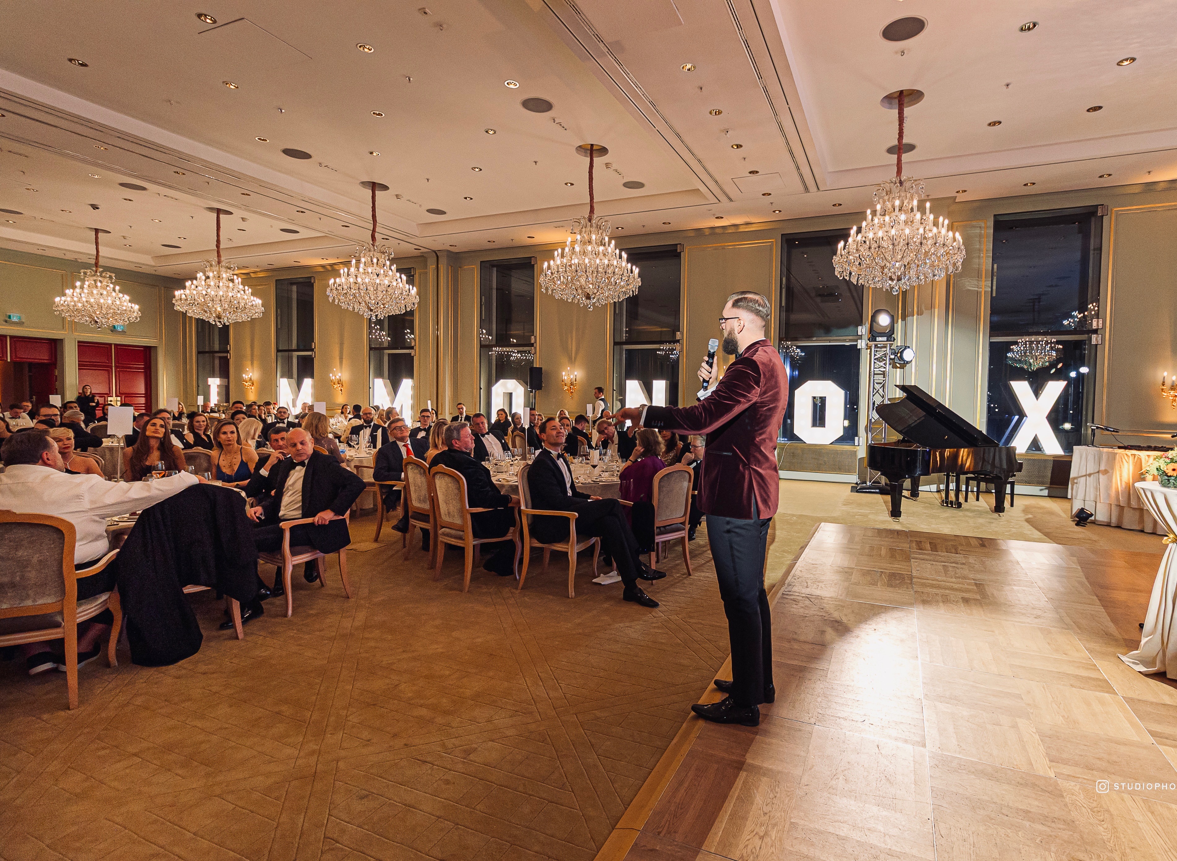 Ein Mann mit Mikrofon spricht zu Gästen bei einem festlichen Abendessen in einem eleganten Saal.