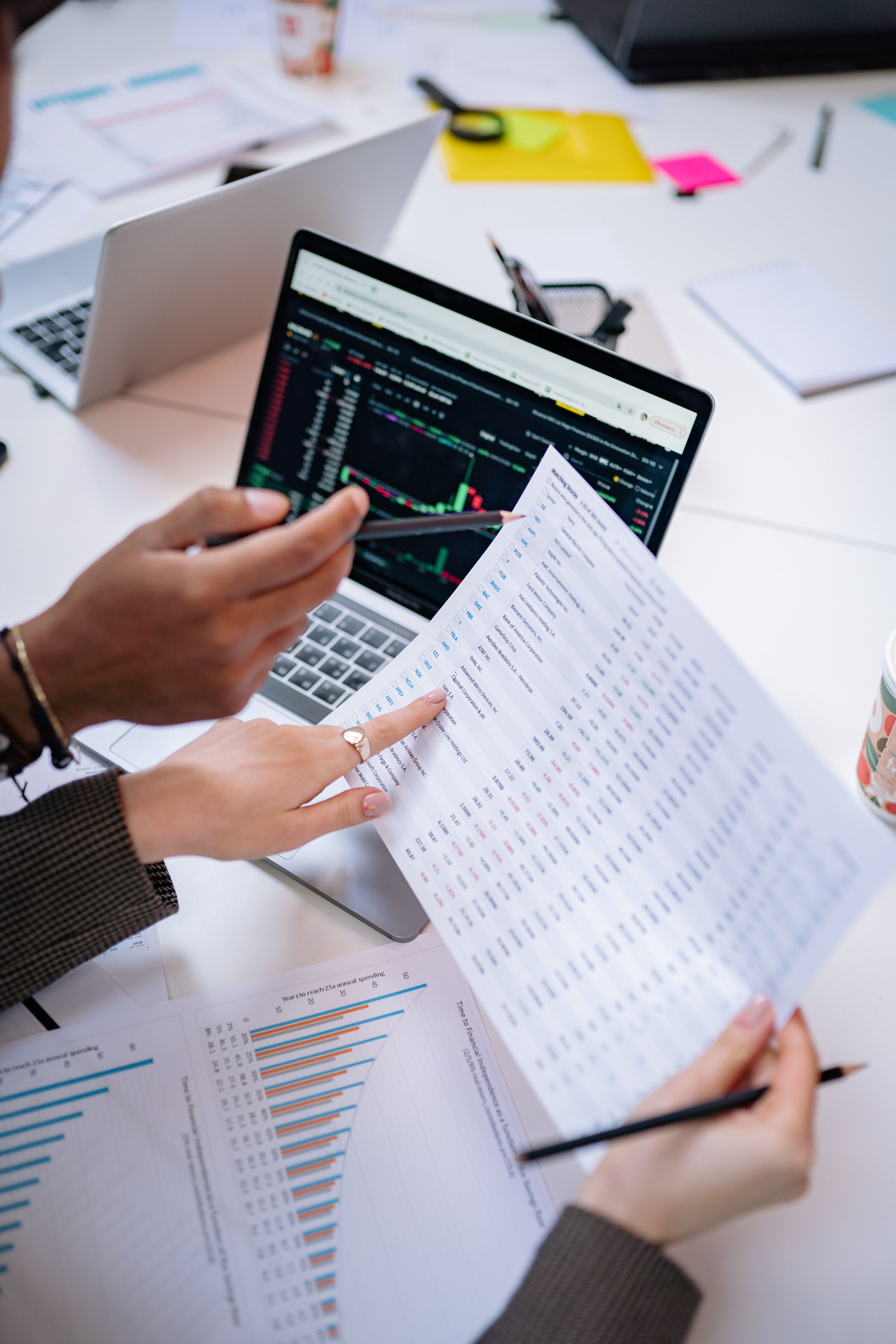 Two people reviewing financial charts and stock data on laptop and printed paper.