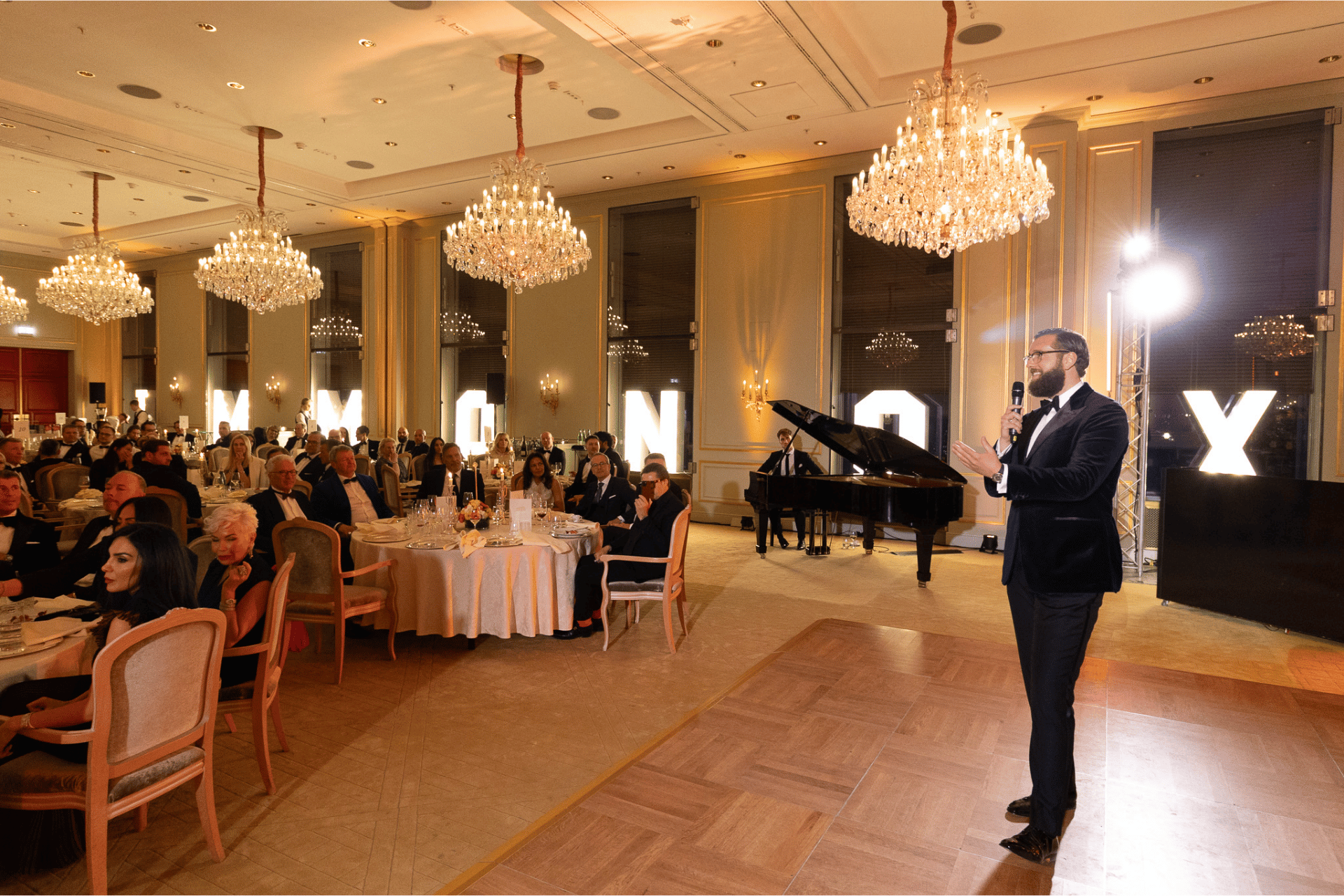 Man in burgundy jacket speaking with microphone to seated audience in an elegant banquet hall.