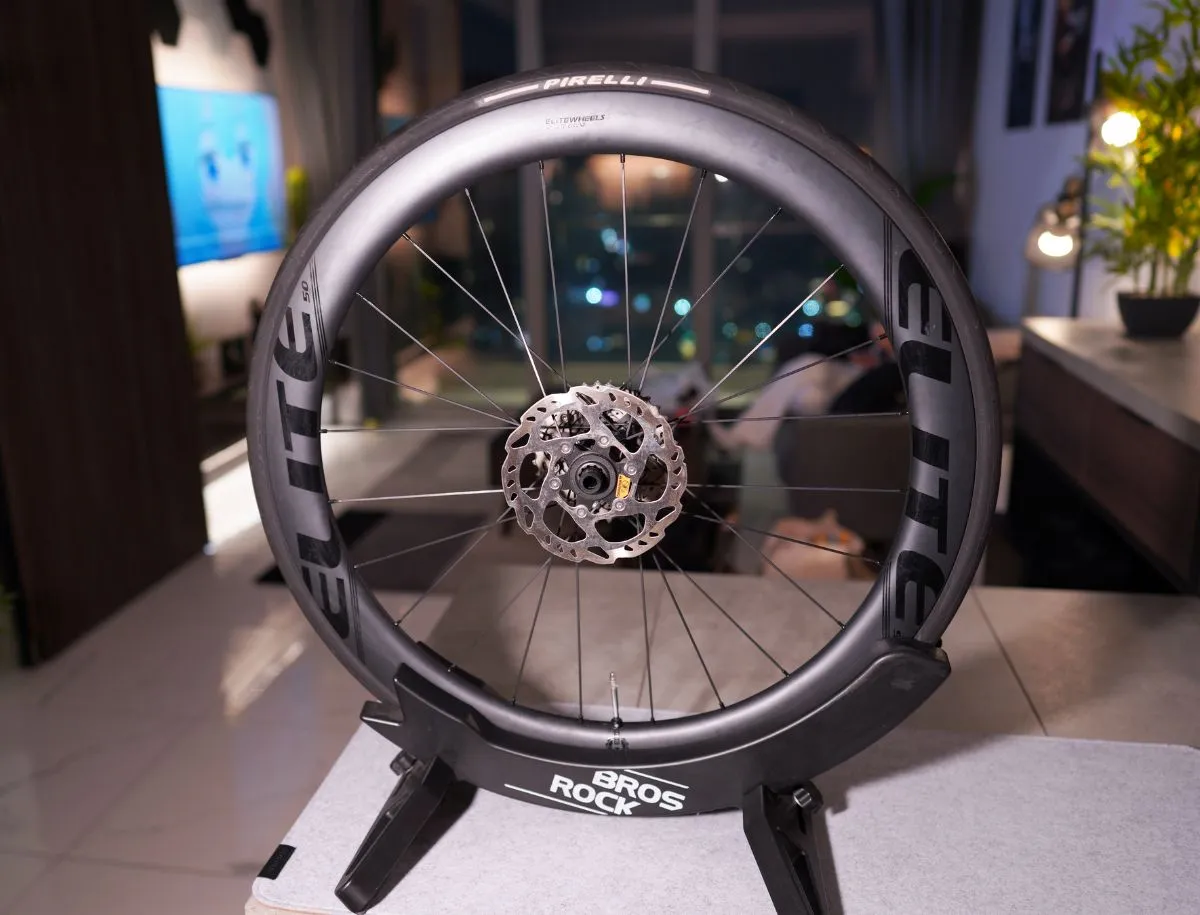 Elitewheels ENT 2.0 50mm carbon road wheel mounted on a display stand with Pirelli tire fitted, disc brake rotor and spokes visible, photographed indoors at night against a blurred apartment background