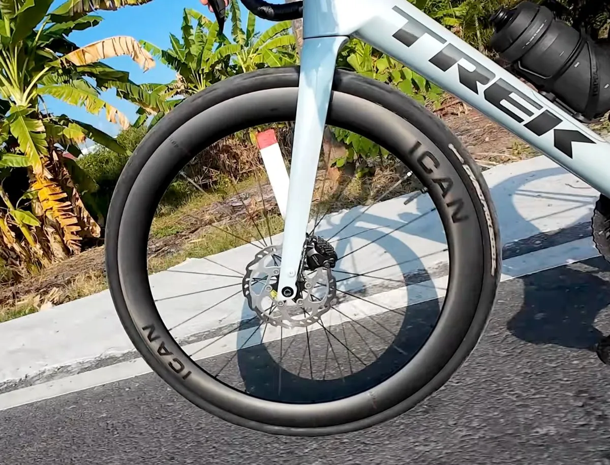 ICAN AERO 50 II carbon front wheel mounted on a Trek Domane SL5, ICAN rim decal visible against a tropical roadside backdrop, disc brake rotor exposed