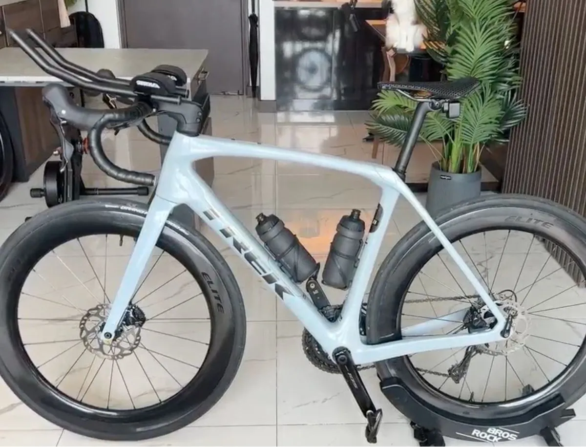 Trek Domane SL5 in Plasma Grey Pearl set up for Ironman triathlon with Profile Design aerobars, Elitewheels deep-section carbon disc brake wheels, three water bottles, and saddle bag, mounted on Bros Rock indoor trainer