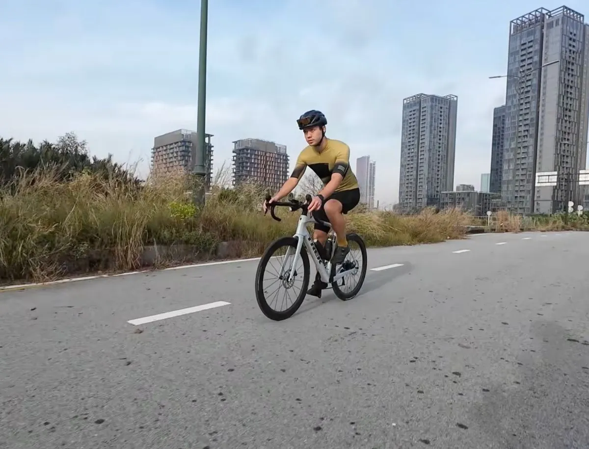 Mike Dee riding a Trek Domane fitted with Elitewheels ENT 2.0 carbon wheels on an open road, with city buildings visible in the background