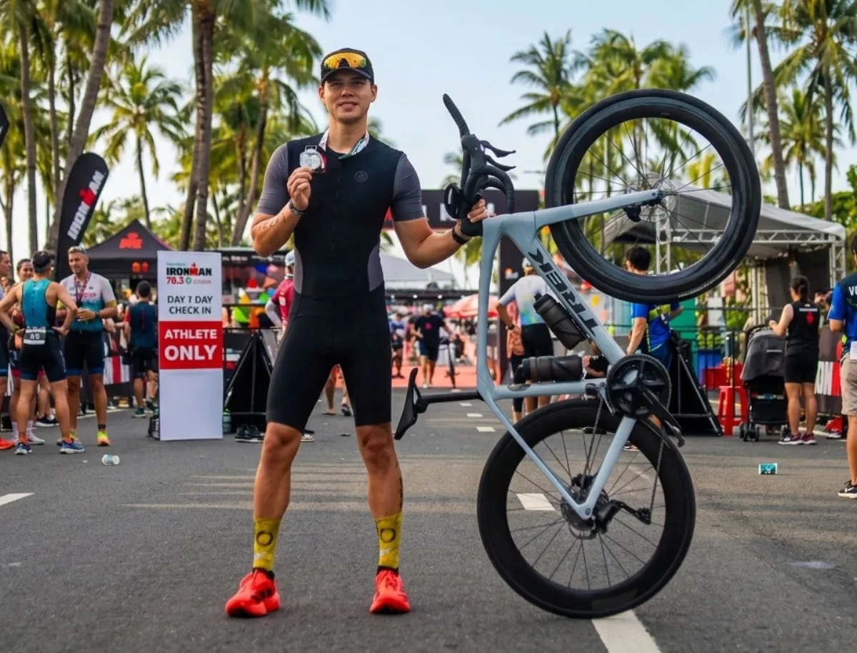 Mike Dee holding an Ironman 70.3 finisher medal at the finish area, standing beside a Trek road bike fitted with Elitewheels ENT 2.0 carbon wheelset
