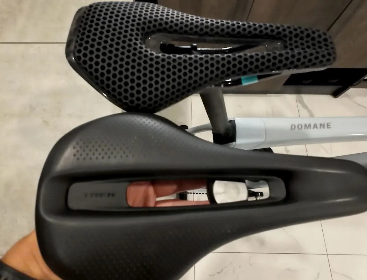 Overhead comparison of two road bike saddles side by side: a Ryet 3D-printed lattice AliExpress saddle on top and a Trek branded saddle with solid foam shell on bottom, Trek Domane frame visible in background