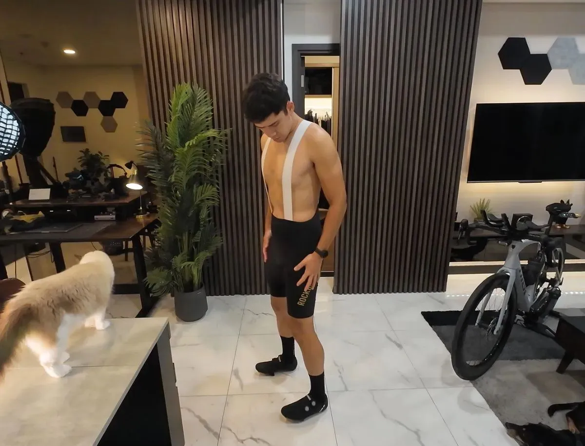 Mike Dee testing black ROCKBROS cycling bib shorts with white straps, checking fit and chamois padding indoors, with a Trek Speed Concept triathlon bike visible in the background for this BikeLabHQ gear review.
