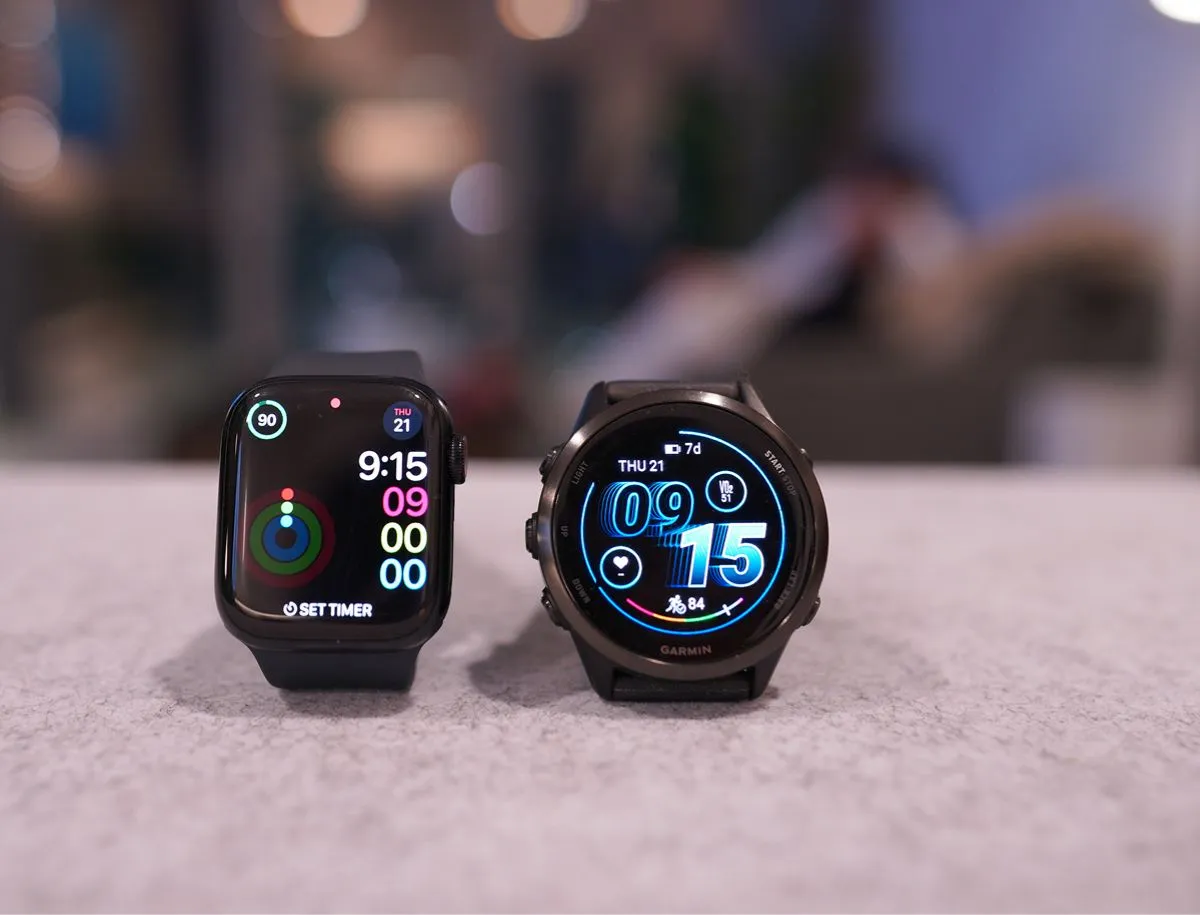 Apple Watch Series 10 and Garmin Forerunner 570 side by side on a gray surface, comparing display size, battery indicators, and heart rate data for cycling and training use
