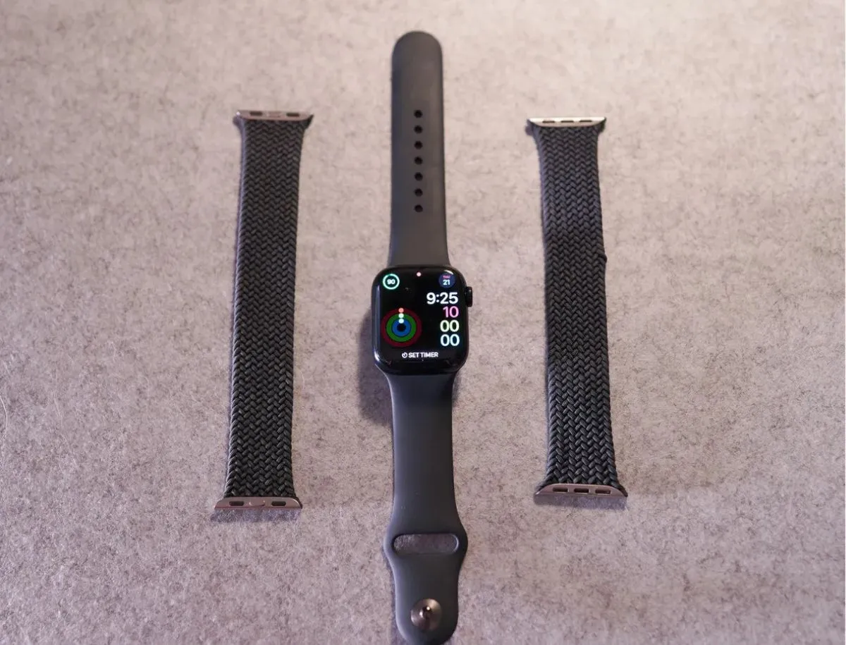 Apple Watch displayed between two black braided Solo Loop bands and a black sport band, laid flat on a gray surface, showing interchangeable strap options.