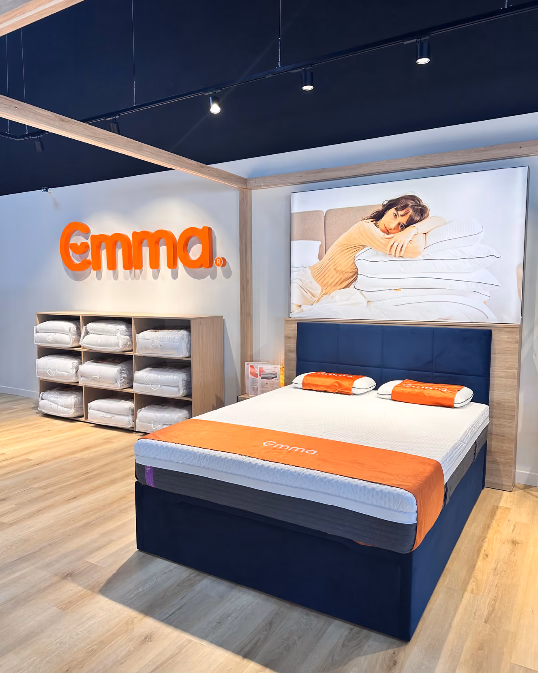 Modern bedroom showroom with Emma mattress, orange pillows, and a large framed photo on wooden floor.