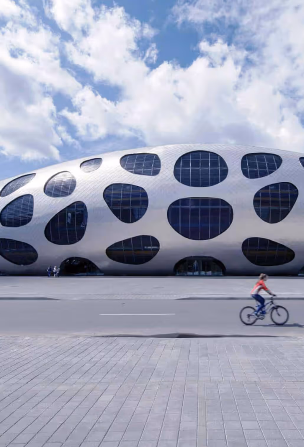 A cyclist rides past a futuristic metallic stadium building featuring a unique, organic pod design and custom-shaped window cutouts.