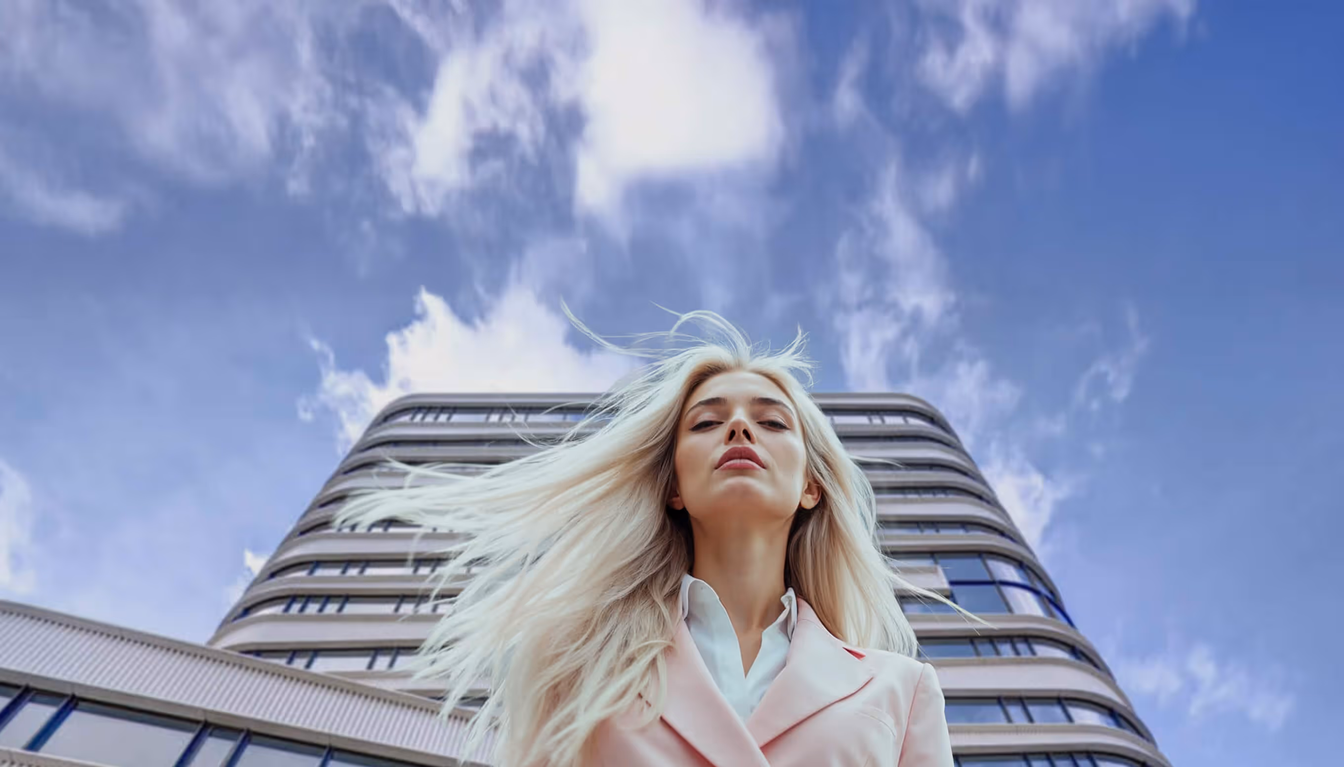 Low angle view of a woman in a pink blazer standing confidently in front of a modern curved architectural building with sleek metal cladding under a cloudy blue sky.
