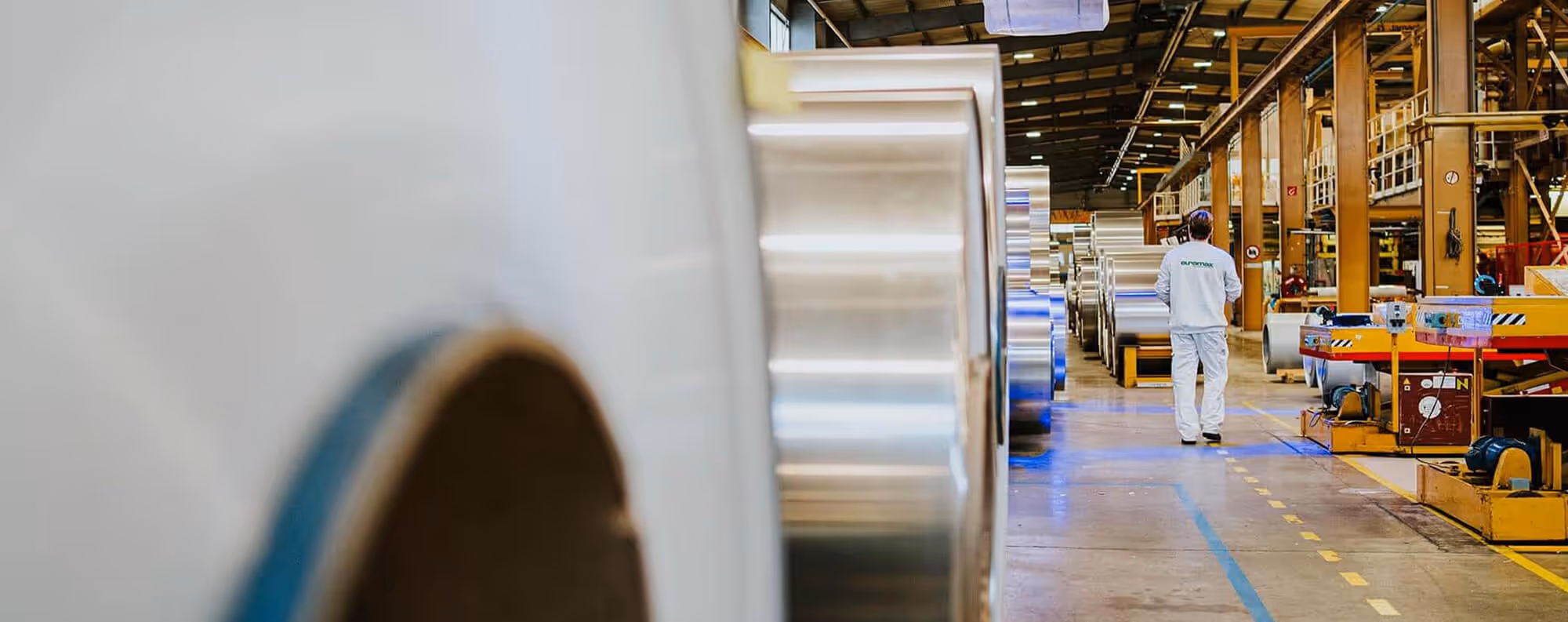 A factory technician walking down an aisle between massive stored rolls of aluminium inside the Euramax coil coating facility.