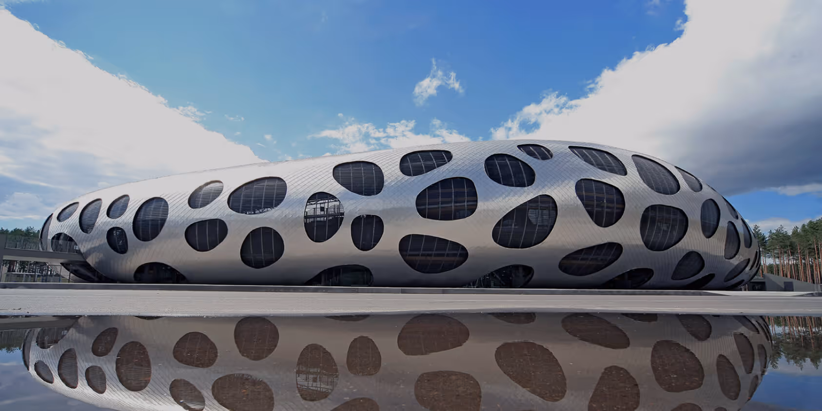 A wide shot of a modern stadium with a curved metallic facade and organic windows, perfectly reflecting in a water puddle under a blue sky.