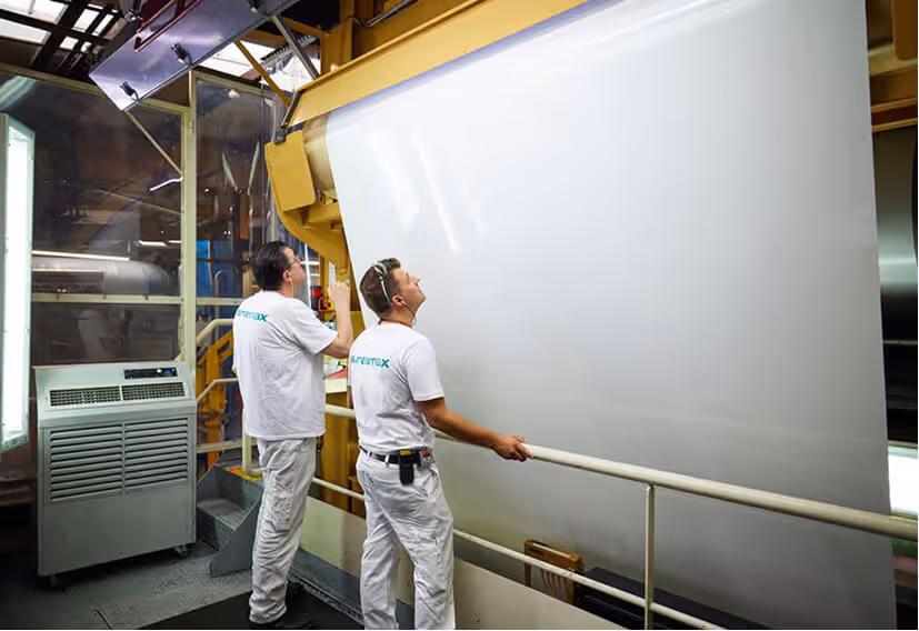Two Euramax production specialists monitor and inspect a large sheet of white pre-coated aluminium moving through the industrial coil coating line.