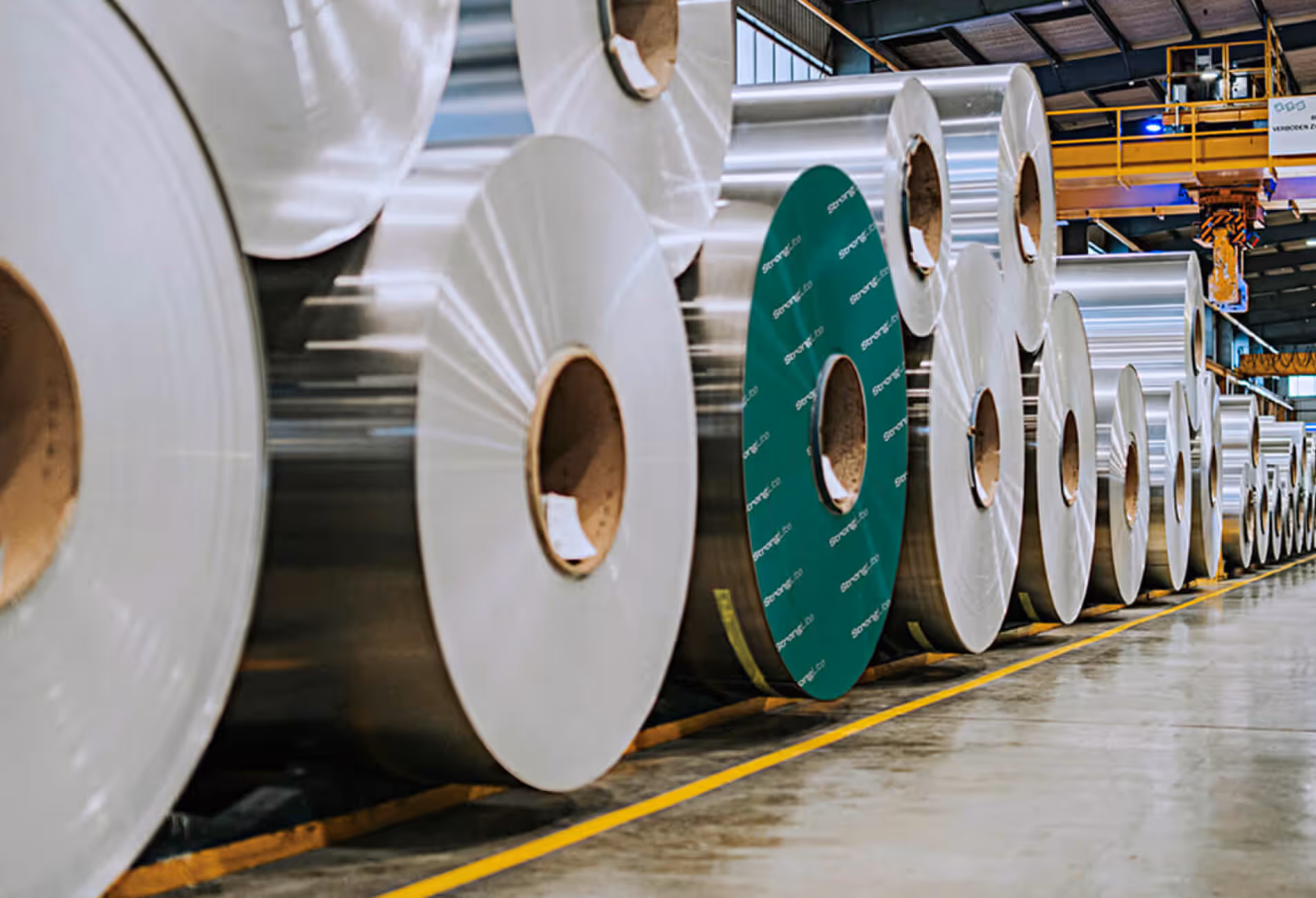 A long row of large, reflective pre-coated aluminium coils lined up inside the Euramax warehouse facility, with one coil featuring a distinct green endcap.