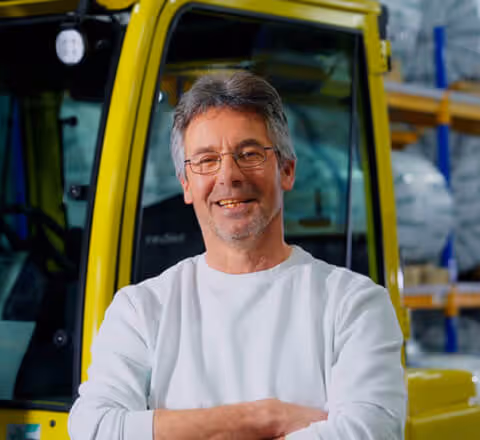 Portrait of a smiling Euramax warehouse team member standing with crossed arms in front of a yellow logistics vehicle.