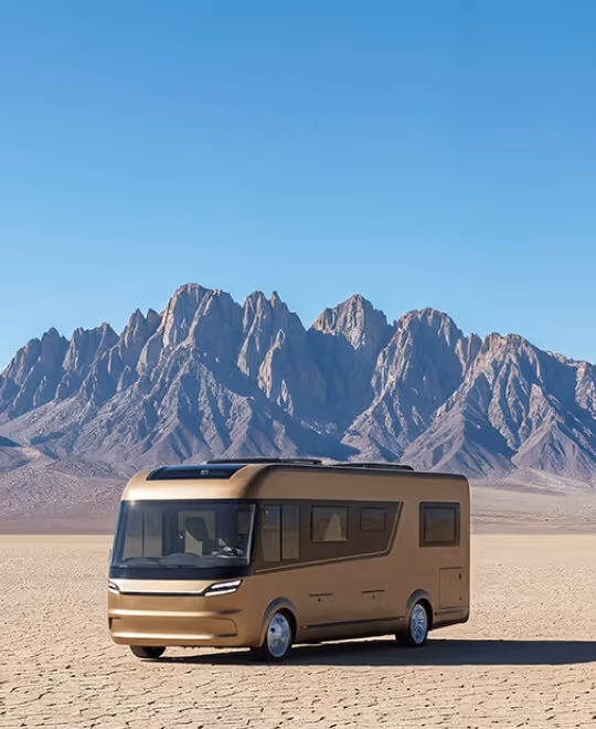 A high-end motorhome with smooth metallic bronze cladding parked in a desert landscape against a rugged mountain backdrop.