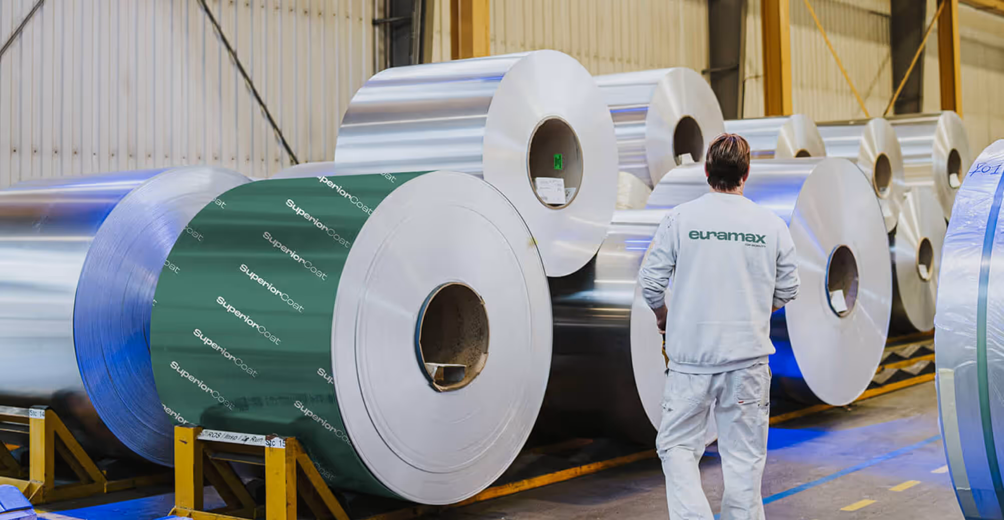 A factory employee walking past large aluminium coils inside a manufacturing facility, highlighting a roll wrapped in a green Euramax SuperiorCoat protective layer.
