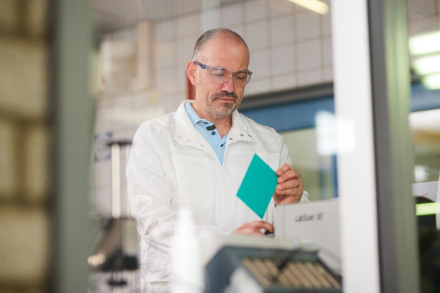A lab technician wearing safety glasses and a white lab coat inspects a teal color sample.