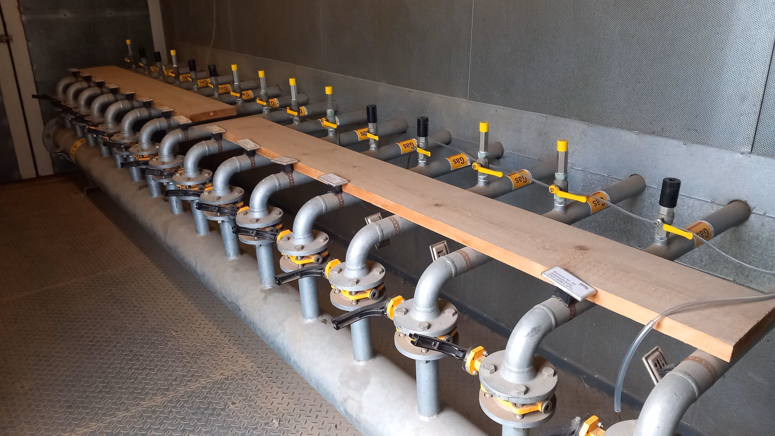 Row of metal gas pipes with yellow-handled valves and labels inside an industrial room.