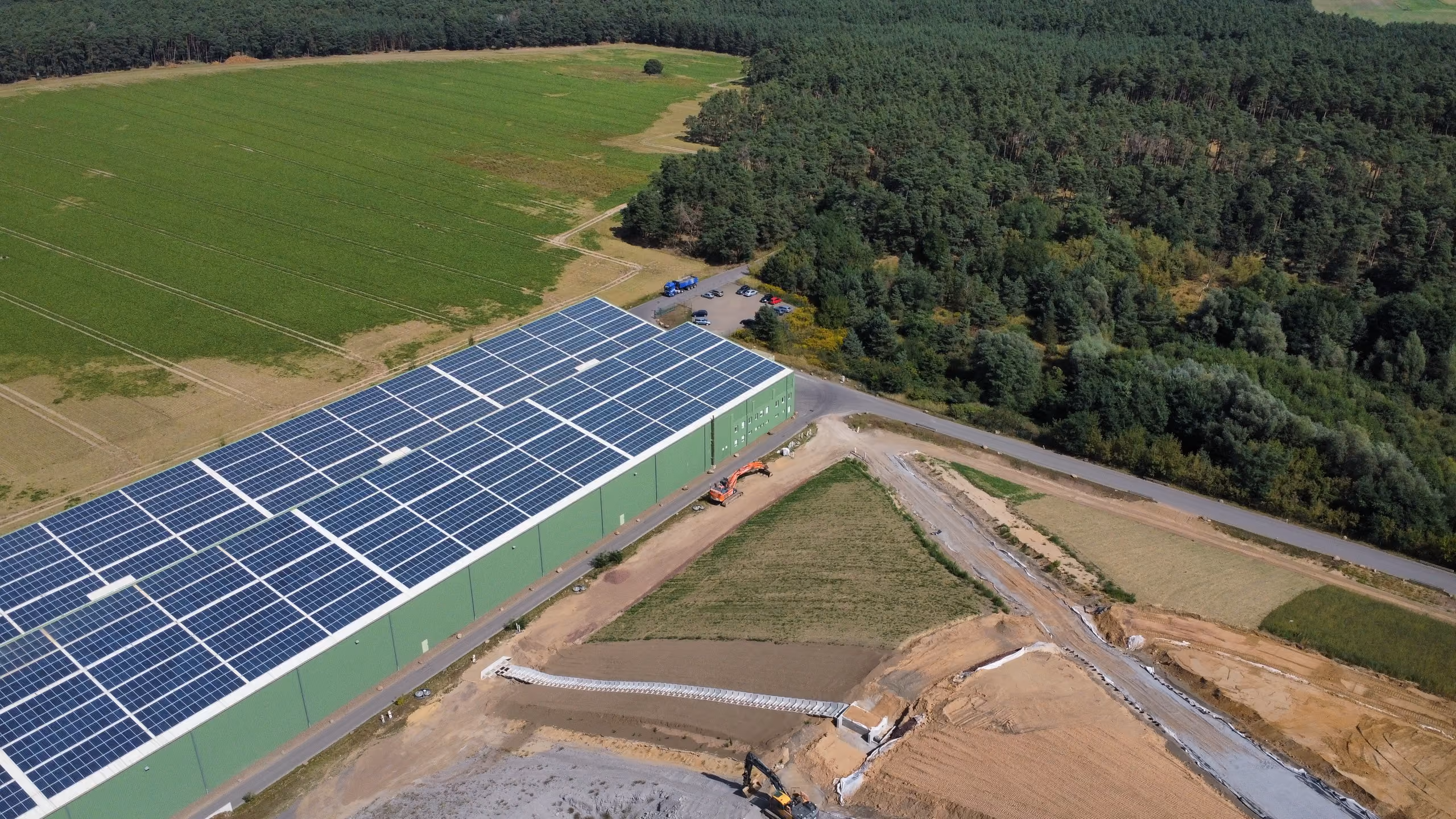 Large green building with solar panels on roof next to forest, fields, and construction site.