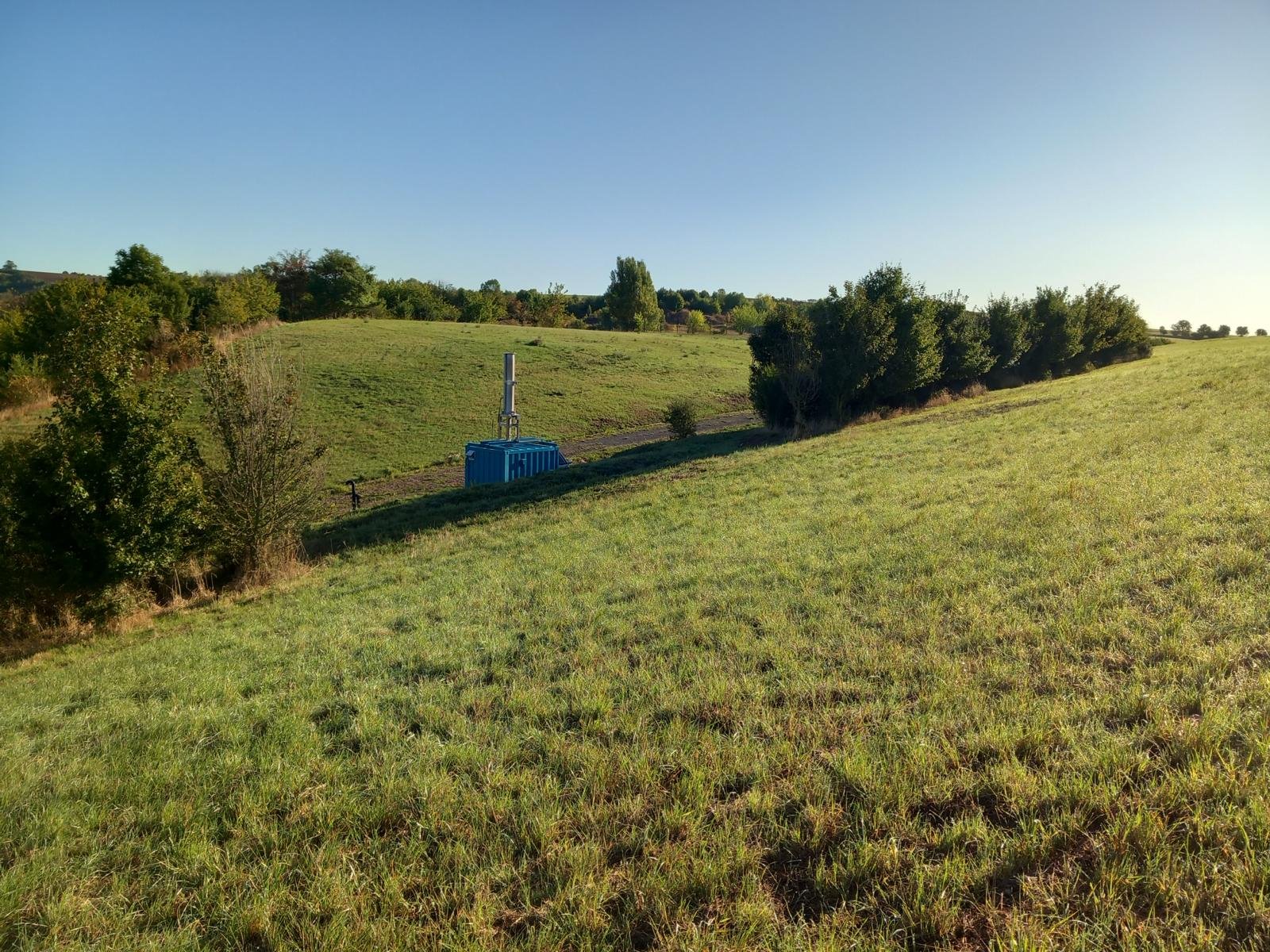 Green grassy field with scattered trees and a blue container with a vertical antenna in the middle.