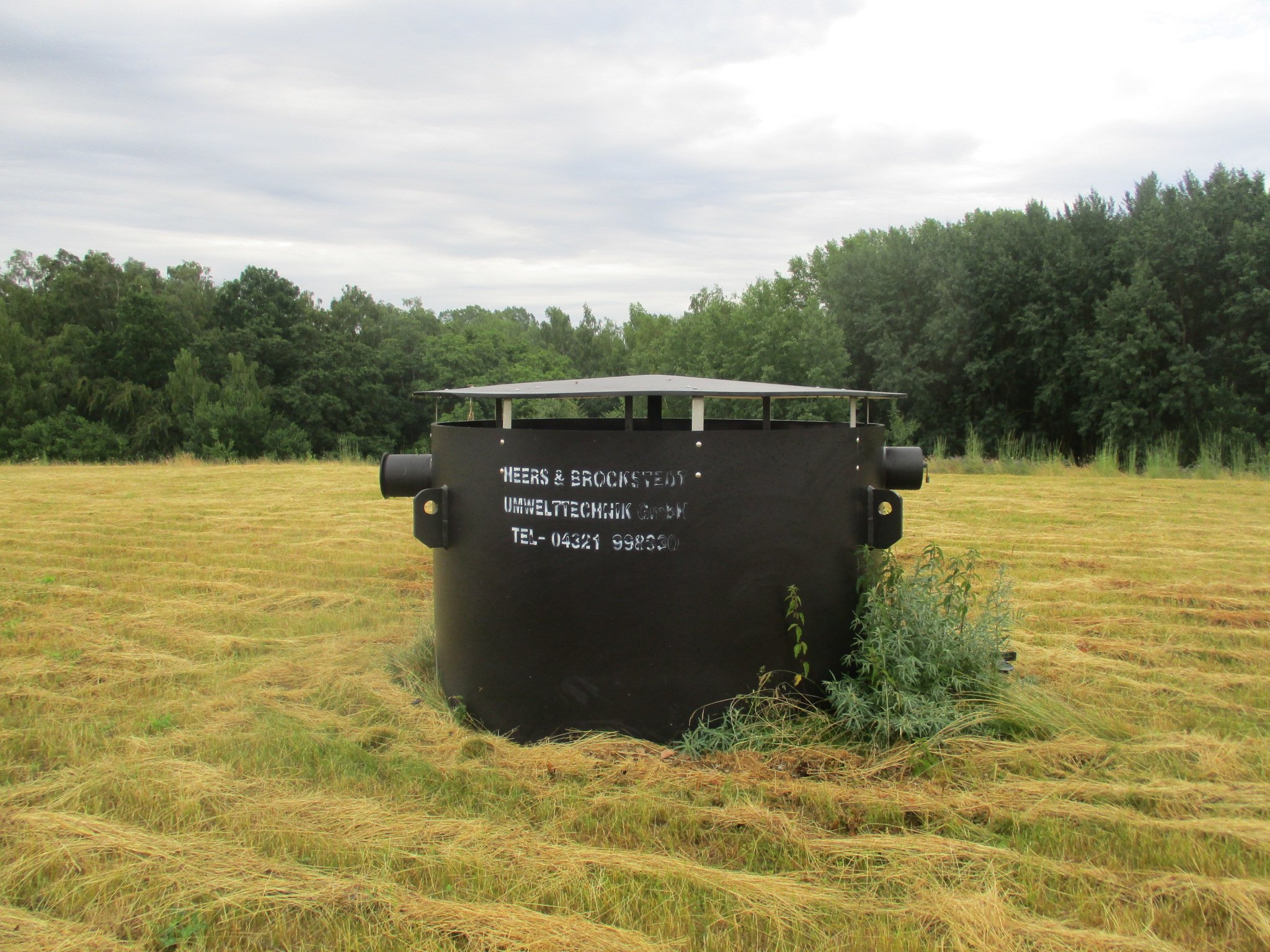 Large black industrial container in a grassy field with trees and cloudy sky in the background.