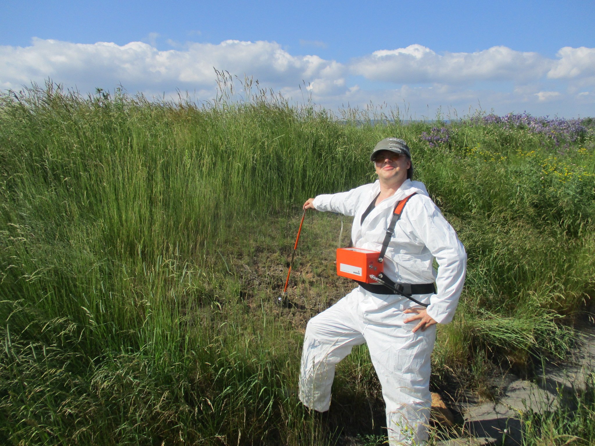 Person in white protective suit and cap holding tool, standing in tall grass under blue sky.