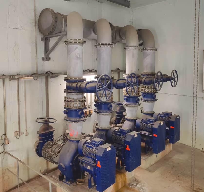 Four large industrial pumps with blue motors and valves connected by thick pipes in a utility room.