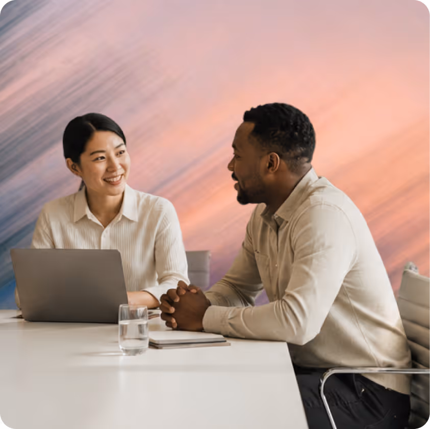 Two coworkers smiling and talking at a desk with a laptop and glass of water.