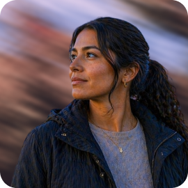 Woman with dark curly hair wearing a jacket looking thoughtfully to the side against blurred background.