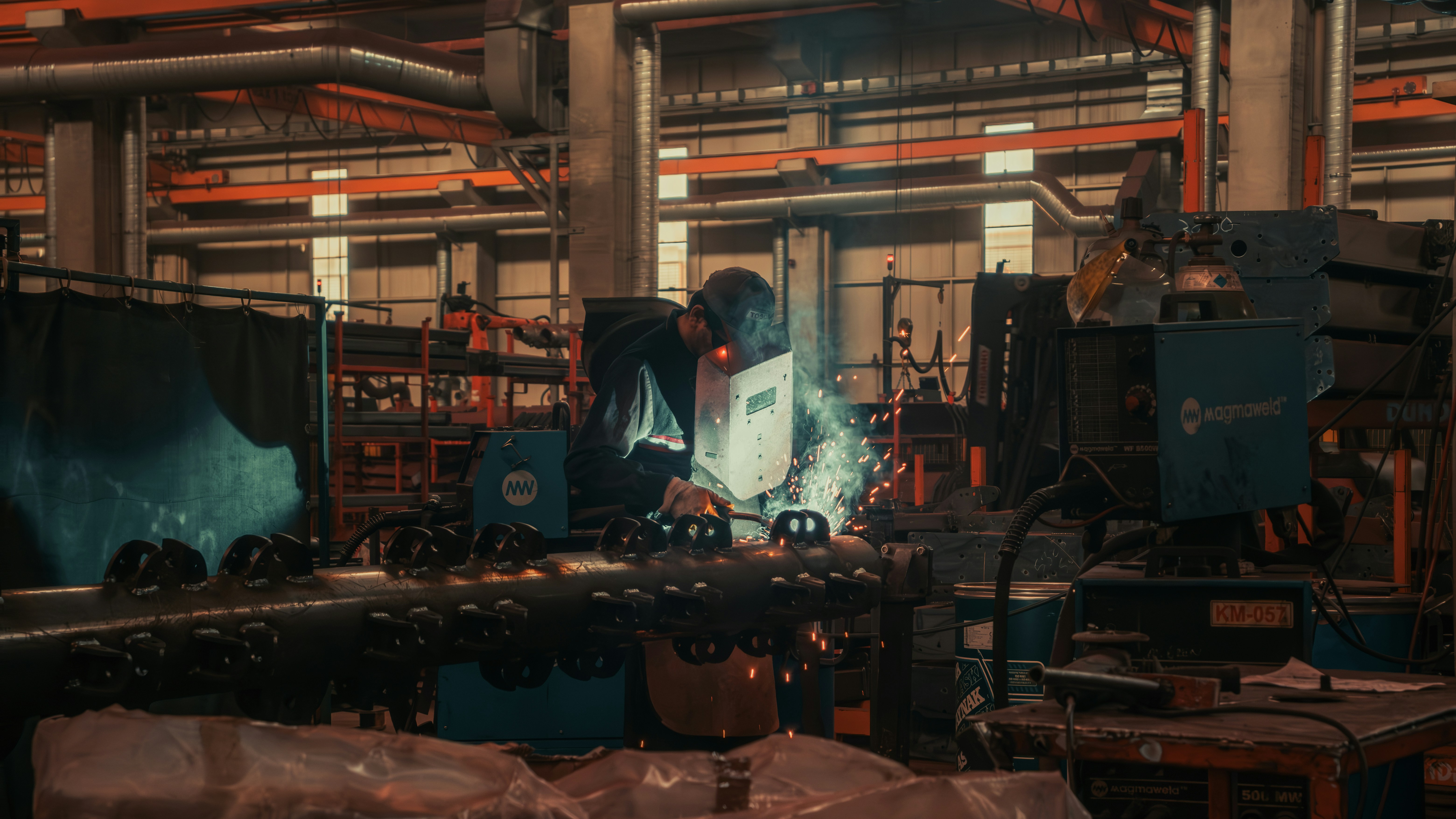 Worker wearing protective gear welding a metal pipe in an industrial workshop with sparks flying.