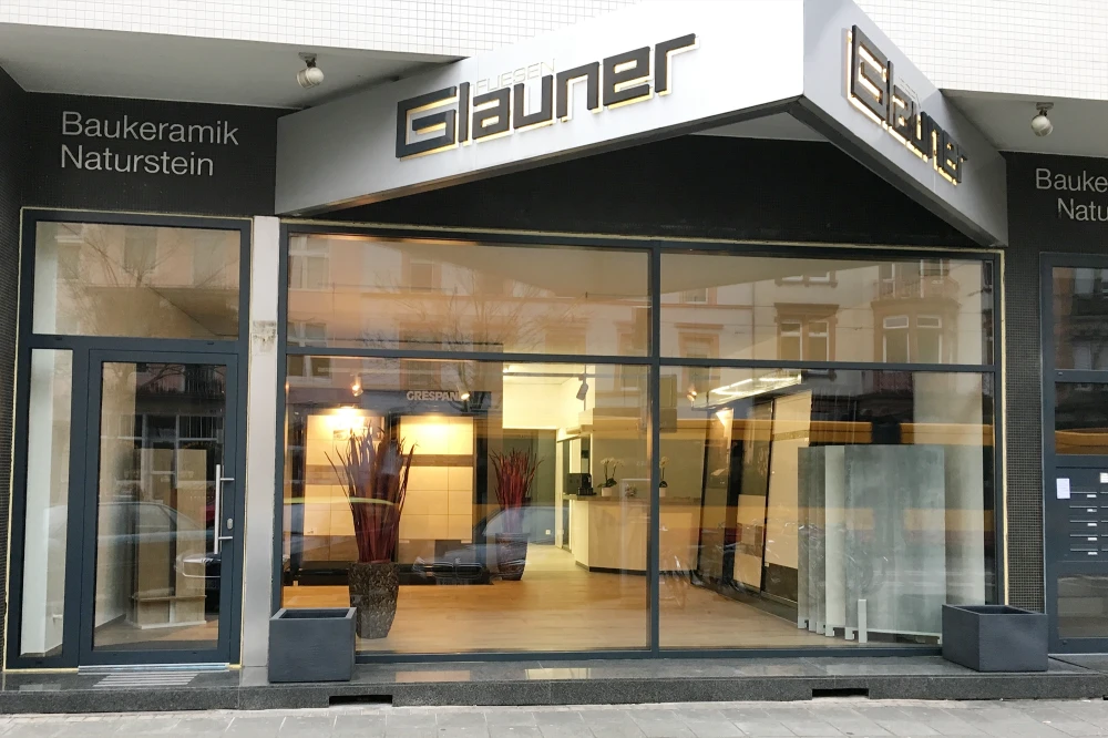 Ladenfront von Fliesen Glauner mit großen Glasfenstern, zwei Pflanzen in Vasen im Inneren und Beschriftung Baukeramik Naturstein außen.