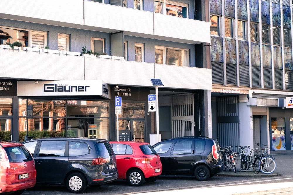 Straßenszene mit mehreren geparkten Autos und Fahrrädern vor einem Gebäude mit dem Geschäftsschild 'Fliesen Glauner', das Fliesen, Baukeramik und Naturstein anbietet.