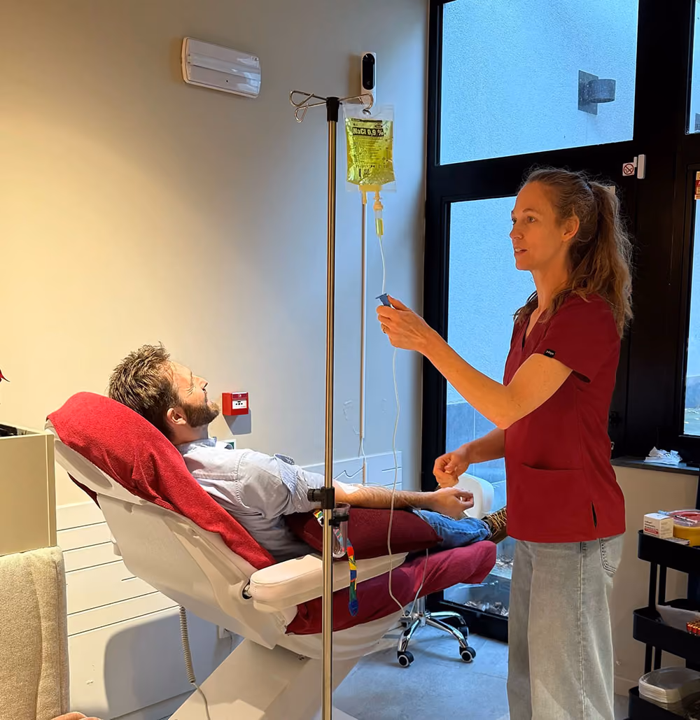 Een patiënt zit ontspannen in een stoel met een infuus, terwijl een zorgverlener het infuus regelt.
