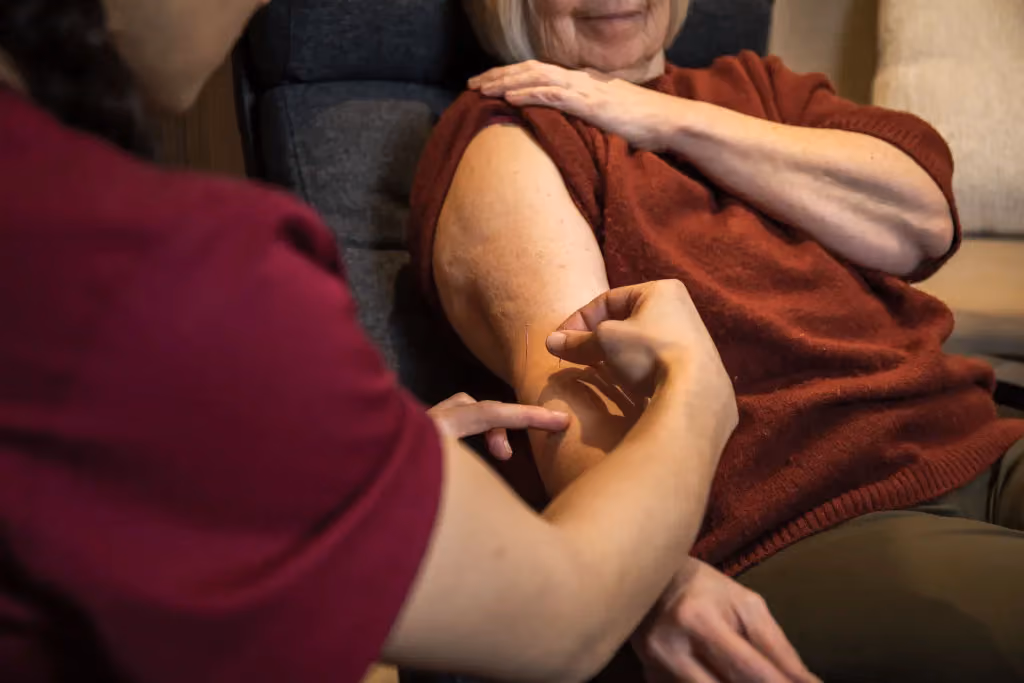 Verpleegkundige die een injectie geeft aan de bovenarm van een oudere vrouw in een rood shirt.
