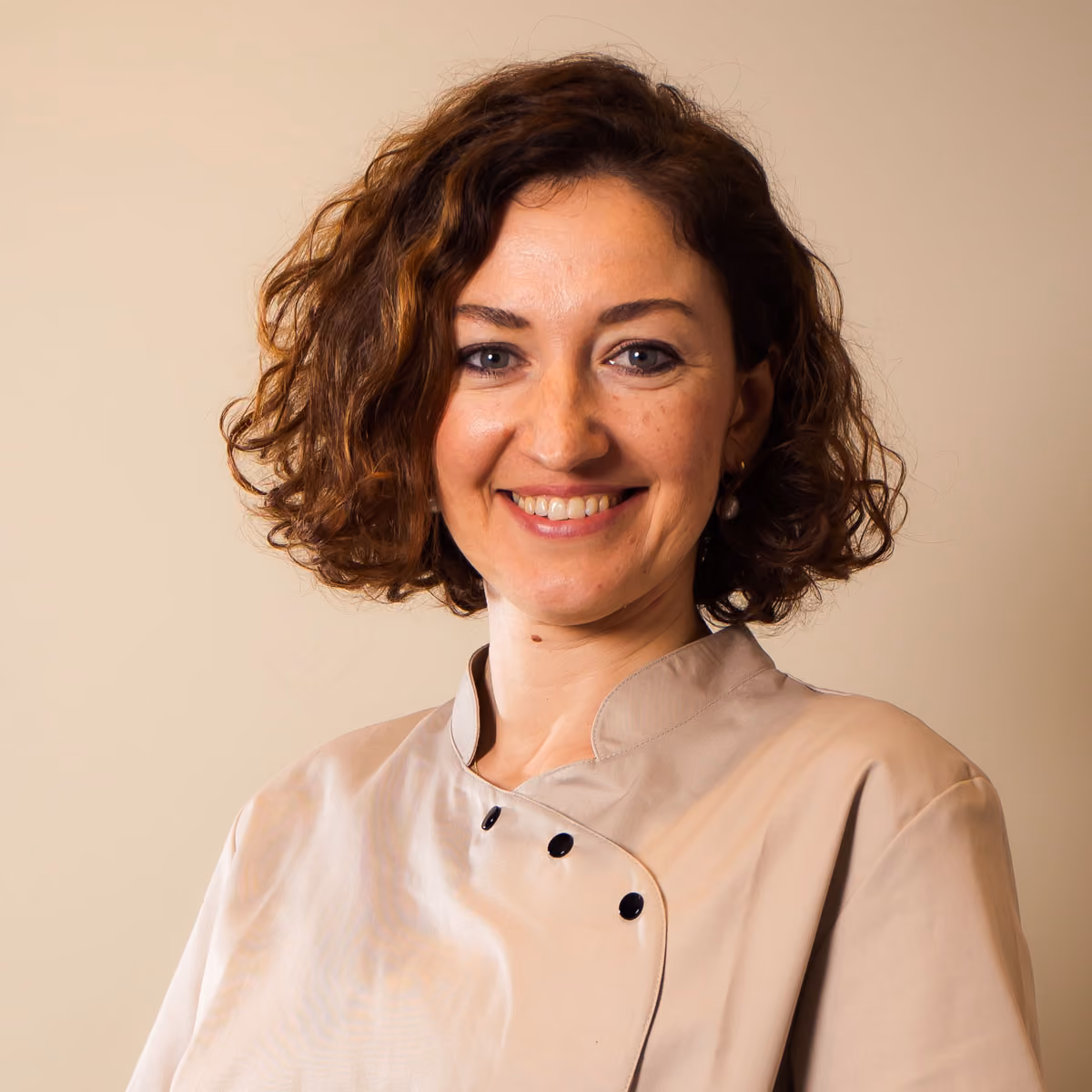 Vrouw met krullend bruin haar en een beige blouse glimlacht tegen een effen achtergrond.
